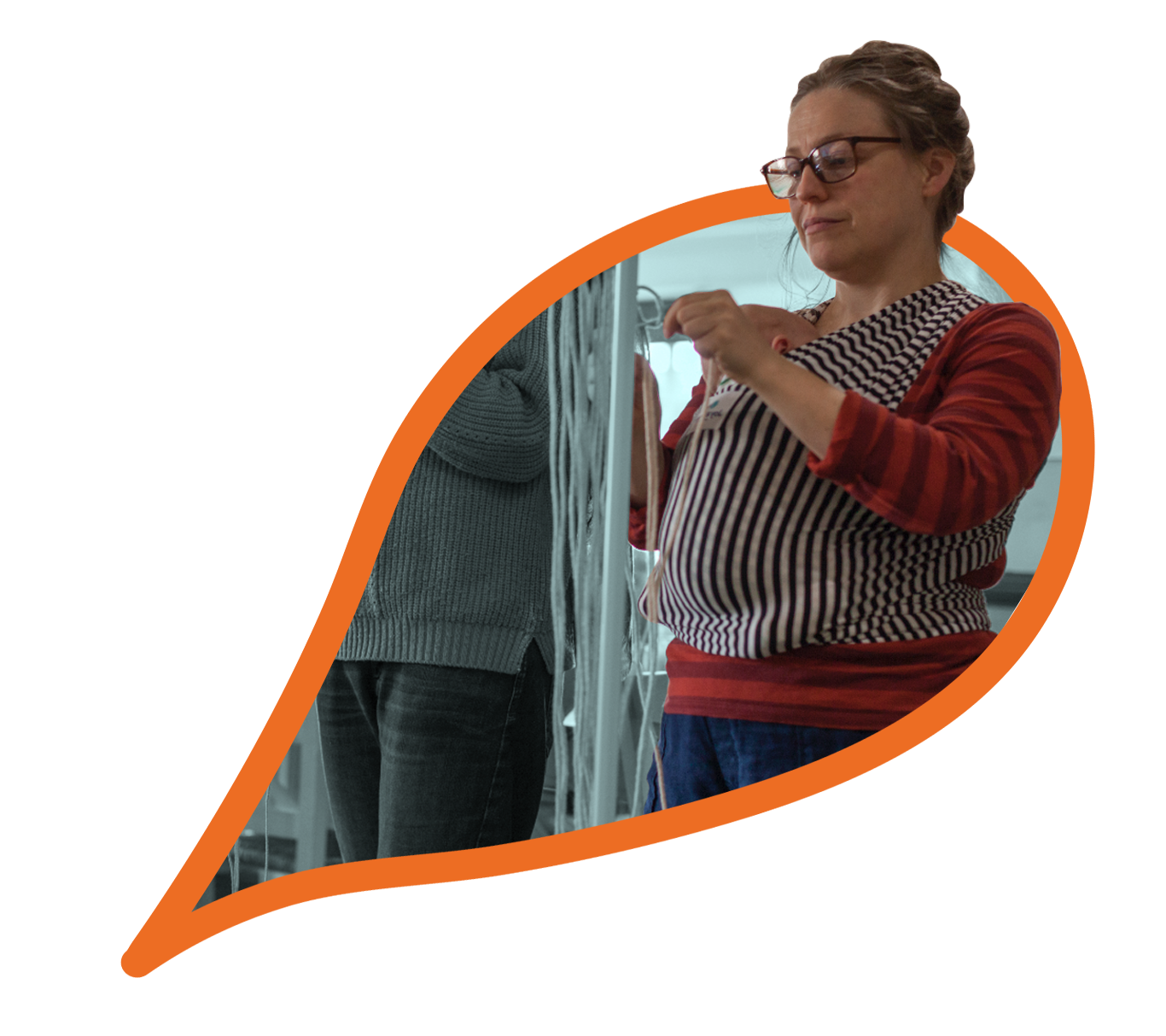 A person with a baby in a sling stands up, knotting and weaving threads in a workshop. 