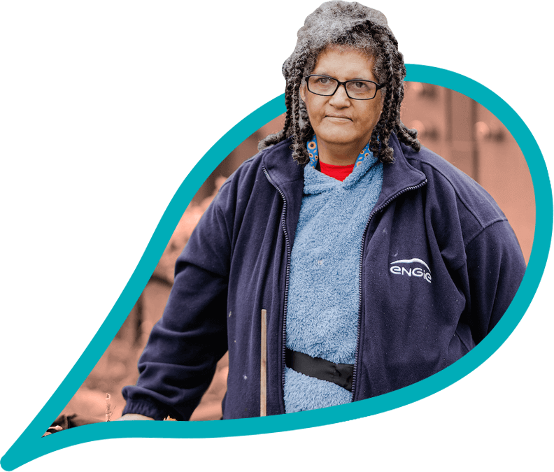 An older person with black braided hair wearing glasses, a sunflower lanyard and a navy fleece stands in a community garden.