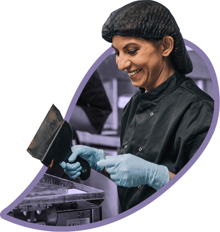 Social entrepreneur Mona Shah smiles, making chocolates in a commercial kitchen. wearing blue vinyl gloves, a black chef jacket and a hair net.