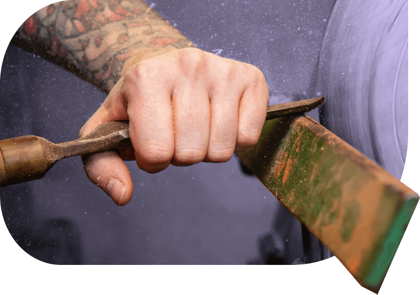 Close up of a hand using a metal tool to shape a long piece of wood against a fast-moving mechanical wheel.