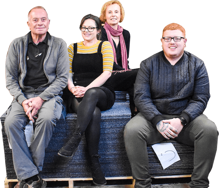 Four members of social enterprise Greenstream Flooring sit on a stack on carpet tiles, smiling.